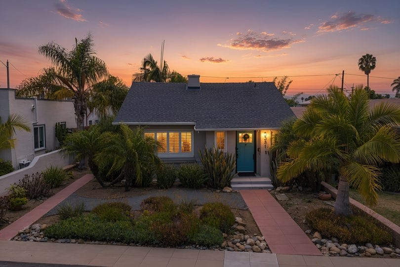 Single-story bungalow home with a grey exterior, turquoise front door, tile walkway, and tropical landscaping at sunset at 3381 Bancroft St in North Park.