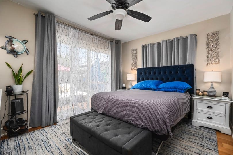 Primary bedroom with blue tufted headboard, dark ceiling fan, sliding glass doors to outdoor deck, white nightstand, and patterned area rug at 3381 Bancroft St in North Park.