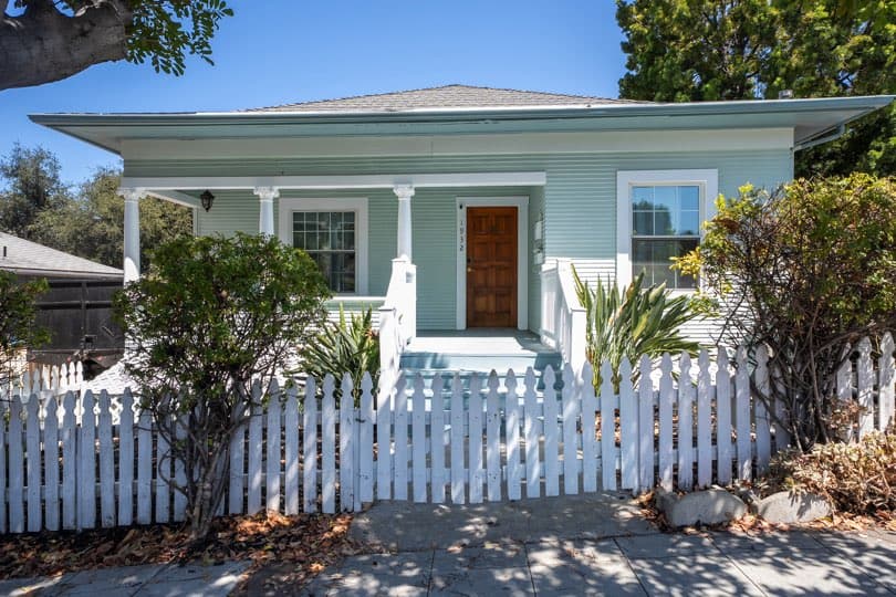 A light mint-green single-story home with a covered front porch and a wooden front door sits behind a white picket fence at 1932 C Street in Golden Hill.