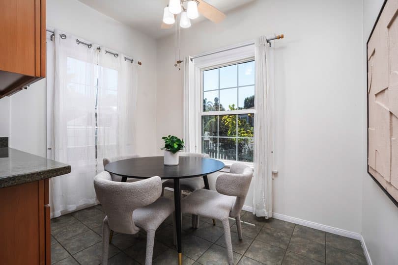 A round black dining table with four grey upholstered chairs is positioned near windows with white curtains at 1932 C Street in Golden Hill.