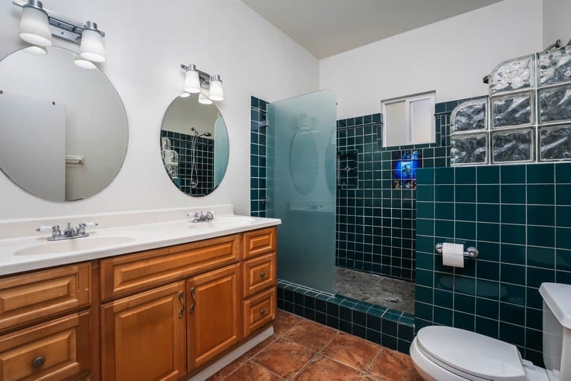 A bathroom features a double-sink vanity with wooden cabinets, dark green tiled walls in the walk-in shower area, a toilet, and round mirrors at 1932 C Street in Golden Hill.