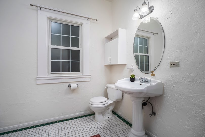 A bathroom features a pedestal sink, a toilet, an oval mirror, and a window with white trim at 1932 C Street in Golden Hill.