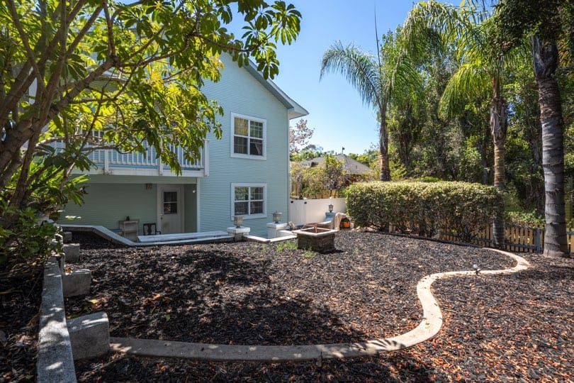 A landscaped backyard with dark mulch and palm trees is viewed next to the light green exterior of 1932 C Street in Golden Hill.