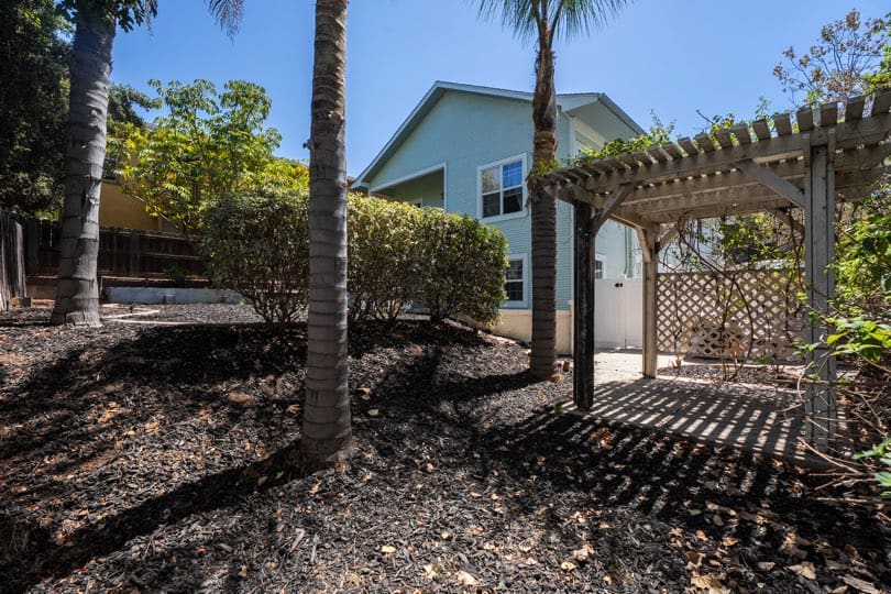 A wooden pergola stands in the backyard with dark mulch and palm trees next to the house at 1932 C Street in Golden Hill.