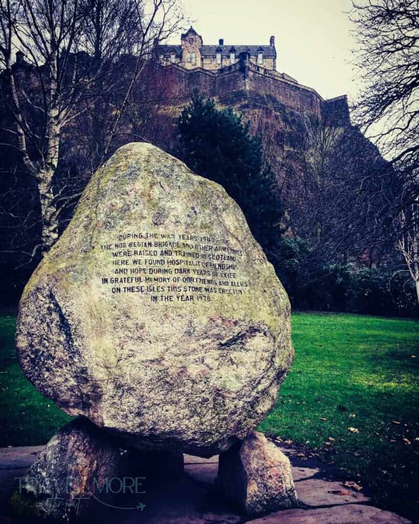 A large inscribed stone stands in the foreground, with a green lawn behind and Edinburgh Castle sitting atop a towering hill above.