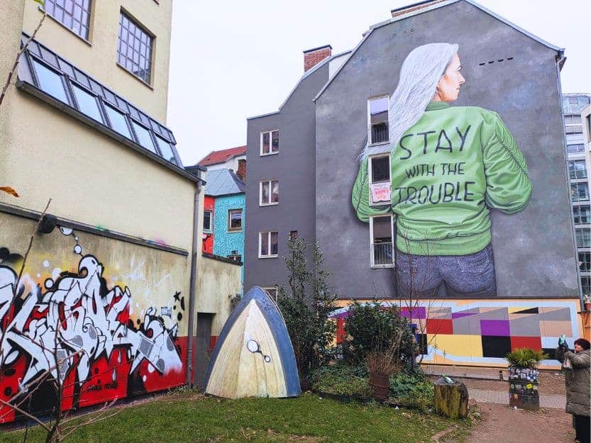 Gängeviertel Street Art in Hamburg Gängeviertel Street Art in Hamburg. Mural of a woman with long grey hair facing forwards wearing a green jacket saying 'STAY WITH THE TROUBLE' on a dark grey painted building