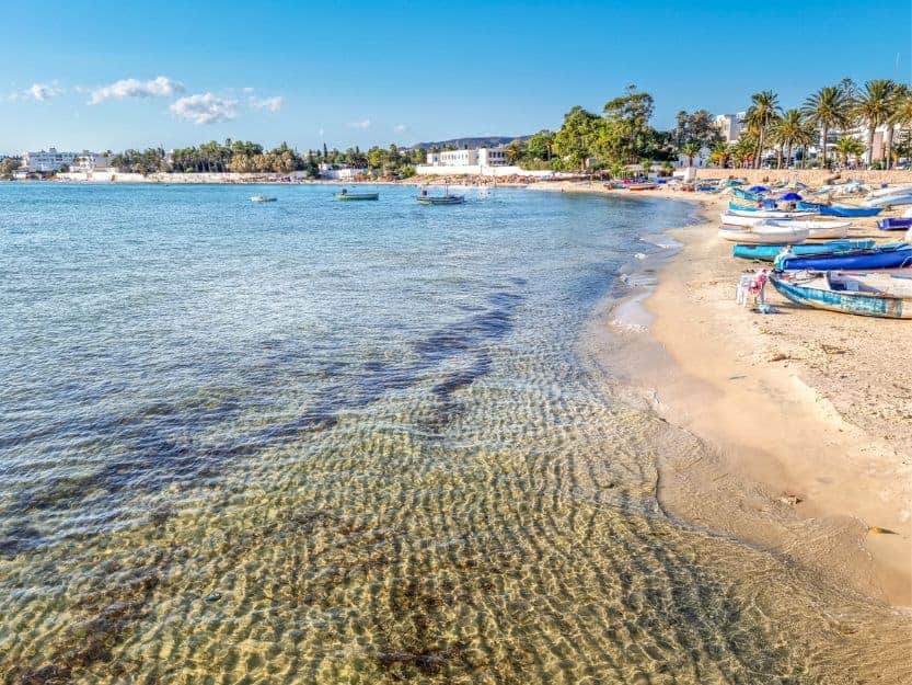 Hammamet beach with golden sand and colourful boats on left and crystal clear bue water on right.