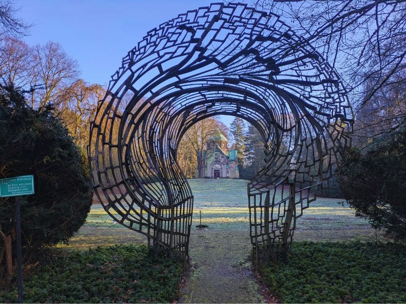 Lost Cemetery of Ohlsdorf in Hamburg Lost Cemetery of Ohlsdorf in Hamburg. Metal sculpture archway with a green roofed mausoleum in the distance.