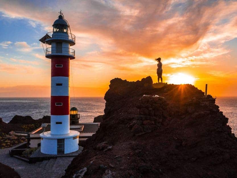 Punta de Teno Lighthouse Punta de Teno Lighthouse. Image of red and white lighthouse at sunset with a woman standing on a rock.