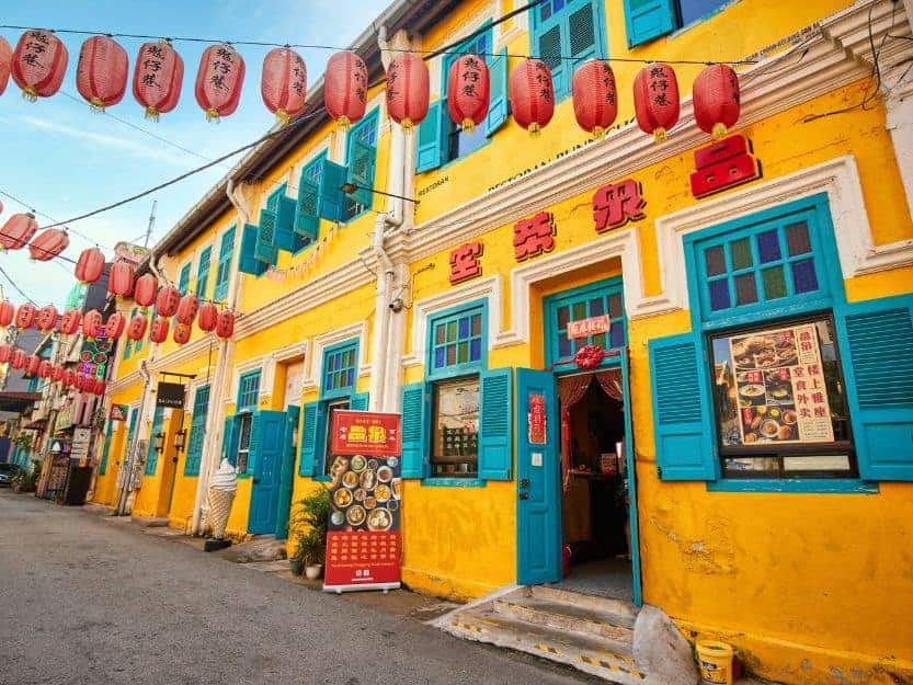 Colourful street in Chinatown in Kuala Lumpur. Yellow buildings with teal doors and shutters with red lanterns strung across the street.