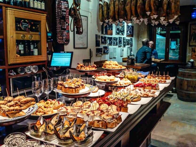 Tapas Bar in San Sebastian in Spain. Lots of tapas dishes in a bar in San Sebastian, Spain. Hams are hanging from the ceiling and two men sitting in the background.