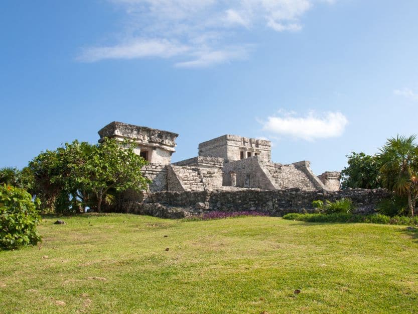 Tulum Ruins in Mexico. Tulums cliffside ruins. Grey stone mayan ruins, surrounded by green palm trees and grass in the foreground. Blue sky with white wispy clouds in the background.