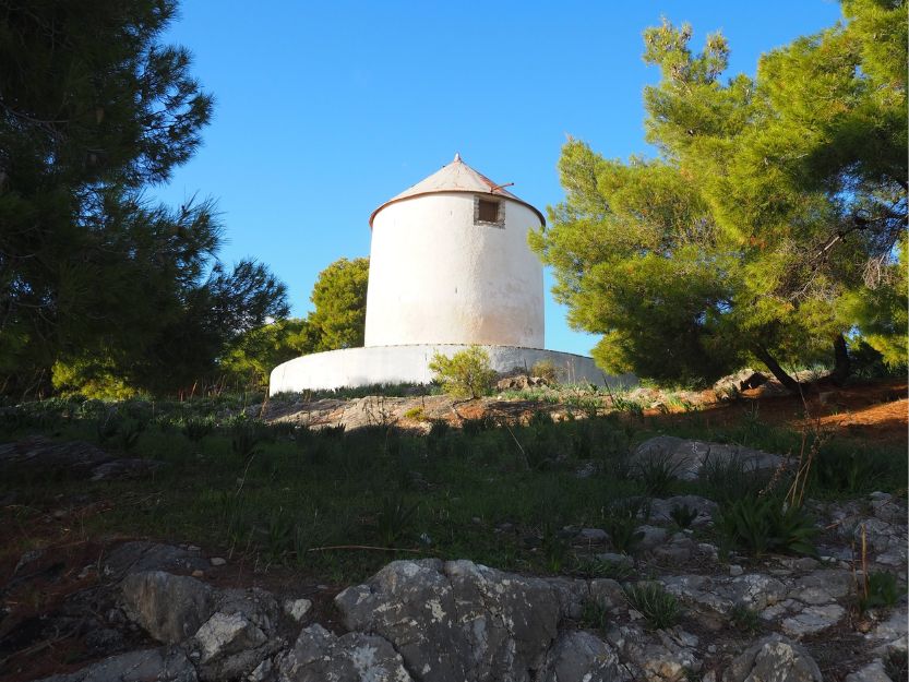 Windmill on the Bisti in Ermioni Greece. Old white windmill on the top of the Bisti in Ermioni. Surrounded by green pine trees with rocks in the foreground and blue sky above