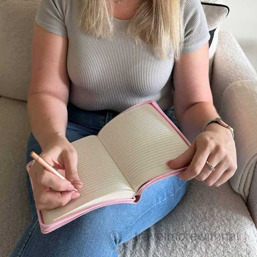 A woman wearing a beige top and blue jeans writes in a journal while sitting on a cream textured sofa. Processing thoughts, successes and learnings is important after travelling with ADHD.