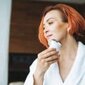 Beauty portrait of smiling woman 35 year in white bathrobe with clean fresh face and hands with red hair doing fasial massage with microcurrent facial massager at the bath room, treat yourself, home body care