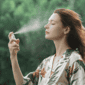 An image of a woman spraying a facial mist on her face on a green background