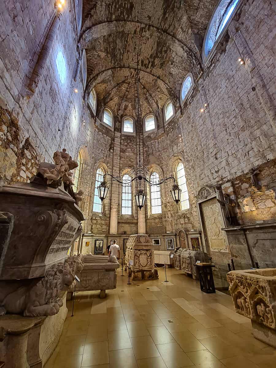 Inside the Carmo Archaeological Museum in Lisbon, the photo displays a collection of ornate sarcophagi set against the rugged stone walls of the Carmo Church, with a visitor in contemplation.