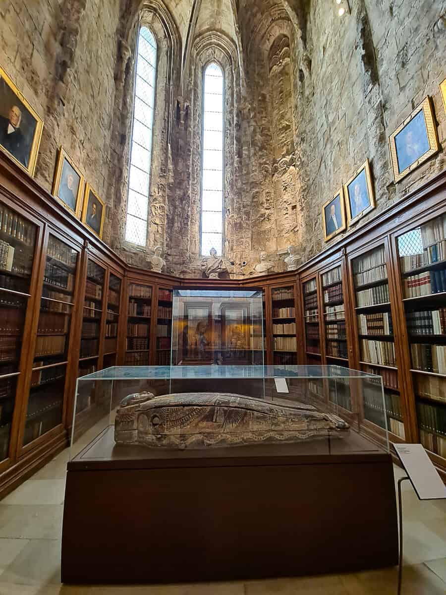 A wide-angle photo of the interior of the Carmo Archaeological Museum in Lisbon, with shelves lined with old books encircling a central glass case displaying an artifact, evoking a sense of historical depth.