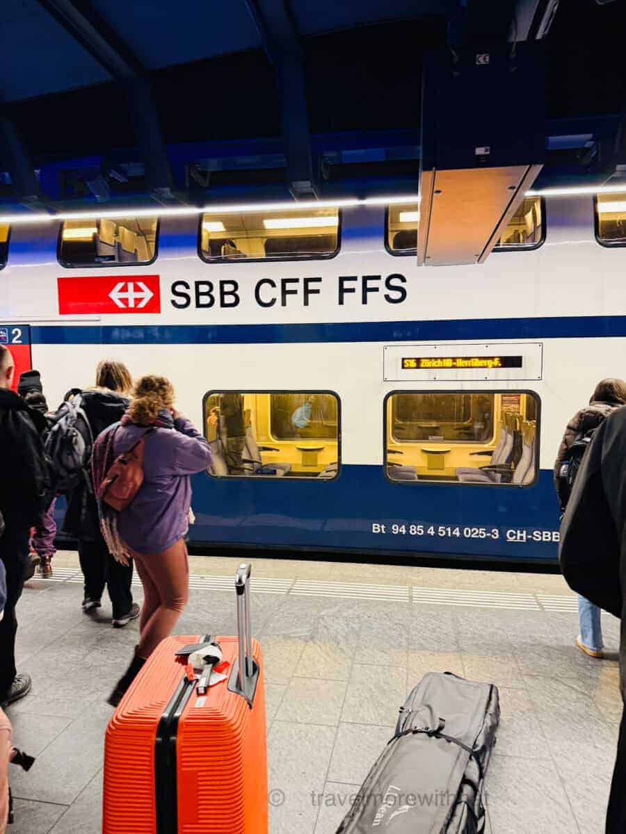 Passengers wait on a train platform, luggage beside them, as a SBB CFF FFS double-decker train pulls in.