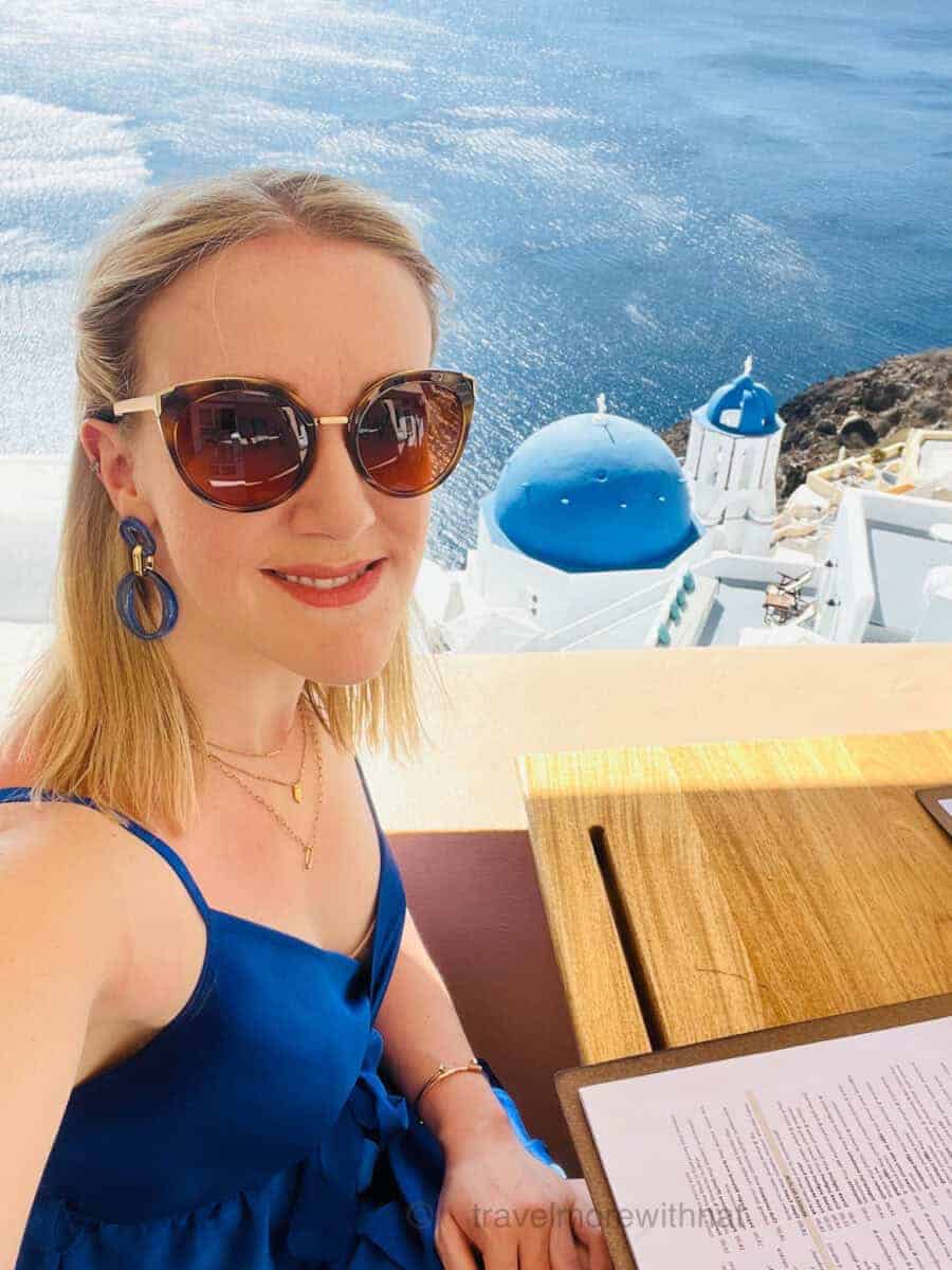 Woman in blue dress, sunglasses, and multiple necklaces, seated at a restaurant table with a menu, overlooking a view of the Aegean Sea and blue-domed buildings in Santorini, Greece. Sometimes finding a cafe with a view is calmer for those travelling with ADHD than battling the crowds at busy sights.