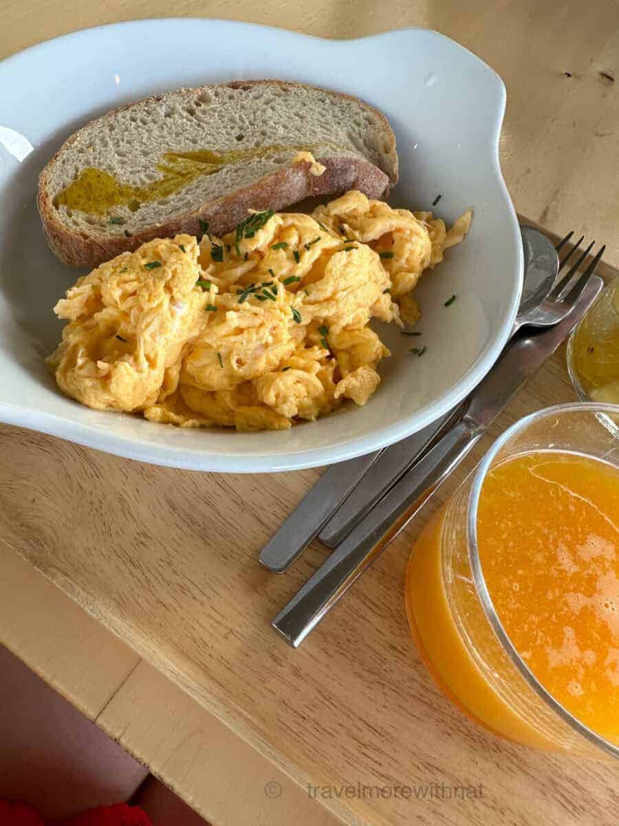 A white bowl of toast and scrambled egg and a glass of orange juice served with cutlery on a wooden table. A good breakfast is important if travelling with ADHD, especially if taking medication.