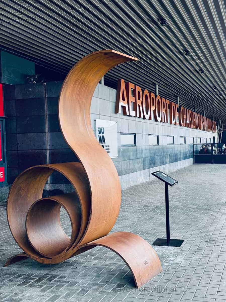 Russet-coloured, curved metal sculpture in front of an airport building with large "AEROPORT DE CHARLEROI BRUXELLES" sign. Special assistance can make navigating airpors easier if travelling with ADHD.