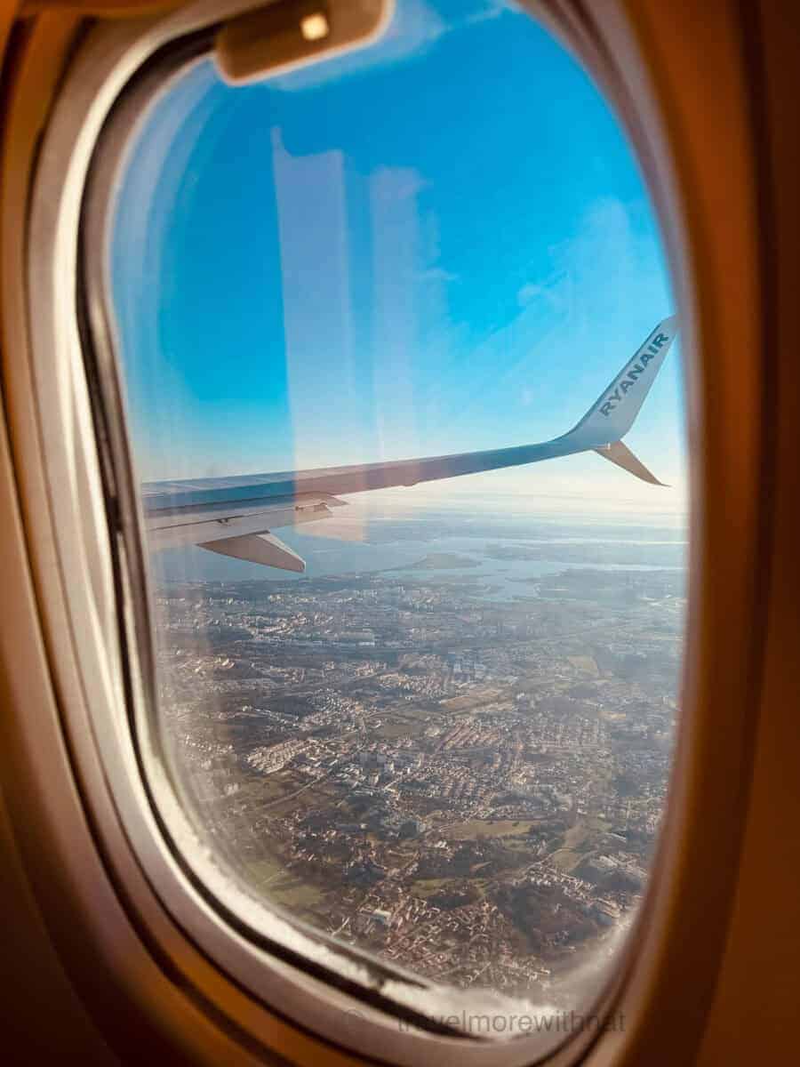 An airplane window view of a coastal city sprawling below, with a portion of the airplane wing and the Ryanair logo visible. A book or game can help pass boredom when flying with ADHD.