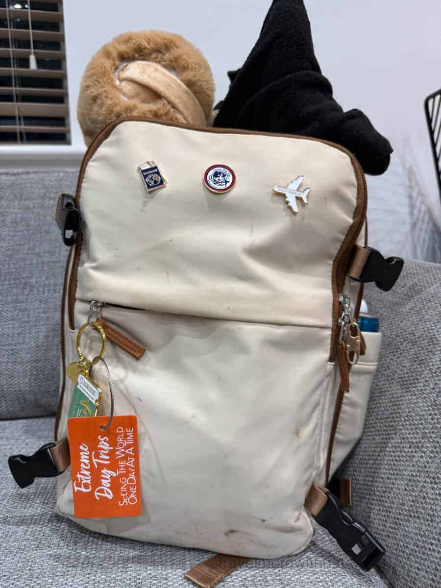 A cream and tan coloured backpack, adorned with pin badges and luggage tags, packed and ready for a trip. Packing early and using a list is essential for neurodiverse travellers.