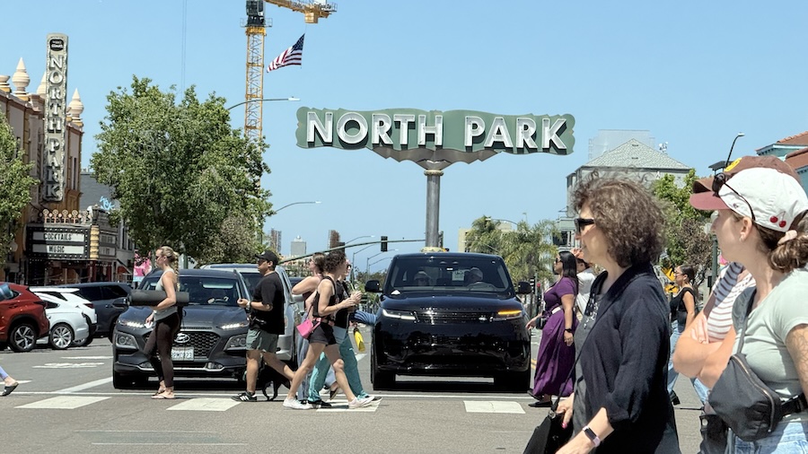 Pedestrians cross University Avenue beneath the North Park sign in San Diego on a sunny day.