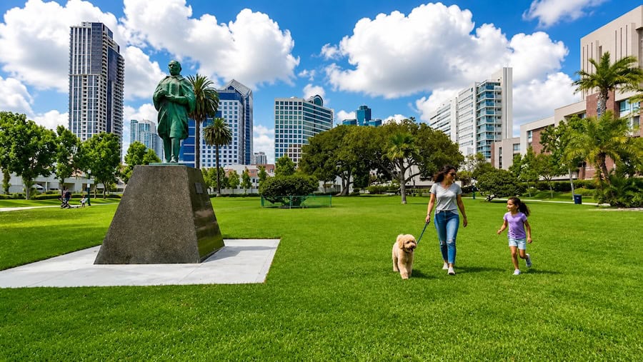 Woman walking a dog with a child in a sunny downtown San Diego park with green grass, puffy clouds, skyline buildings, and a statue in the background.