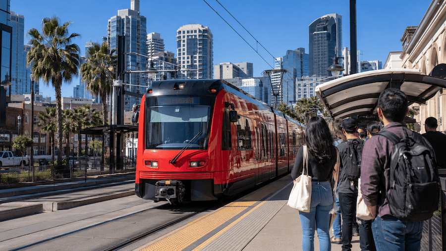 Red San Diego trolley arriving at a downtown station near the Gaslamp Quarter with commuters waiting on the platform and skyline in the background.