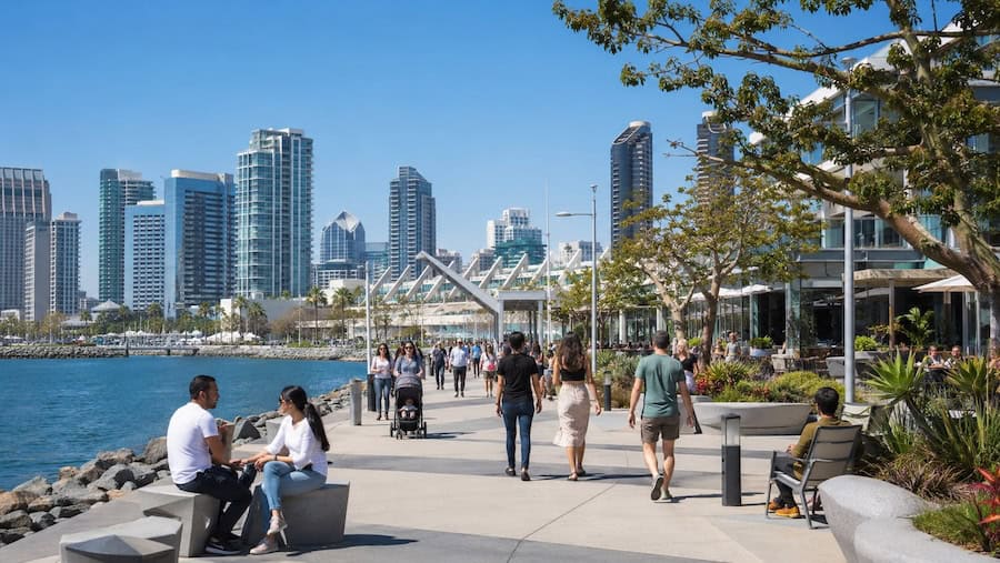 People strolling and relaxing along the Seaport San Diego waterfront promenade with the Downtown San Diego skyline in the background on a bright sunny day.