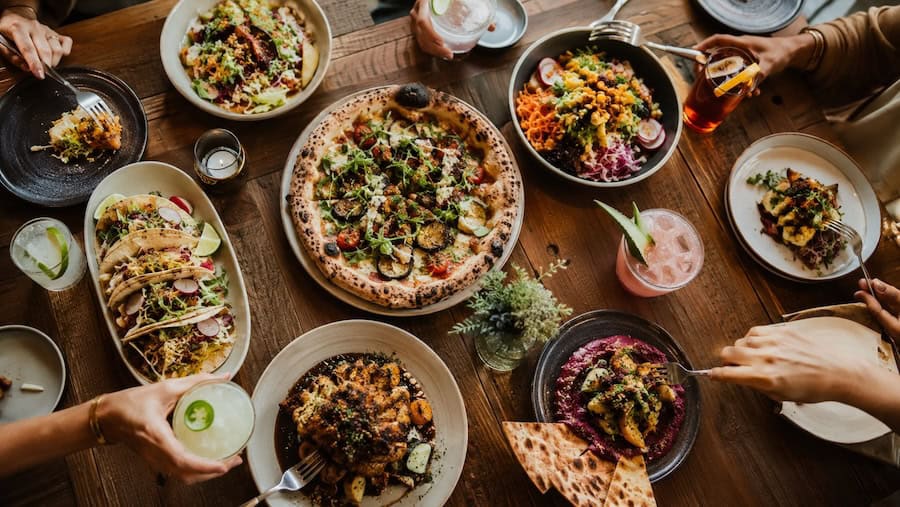 Overhead view of a rustic restaurant table with wood-fired pizza, tacos, vegan dishes, colorful salads, and craft cocktails.