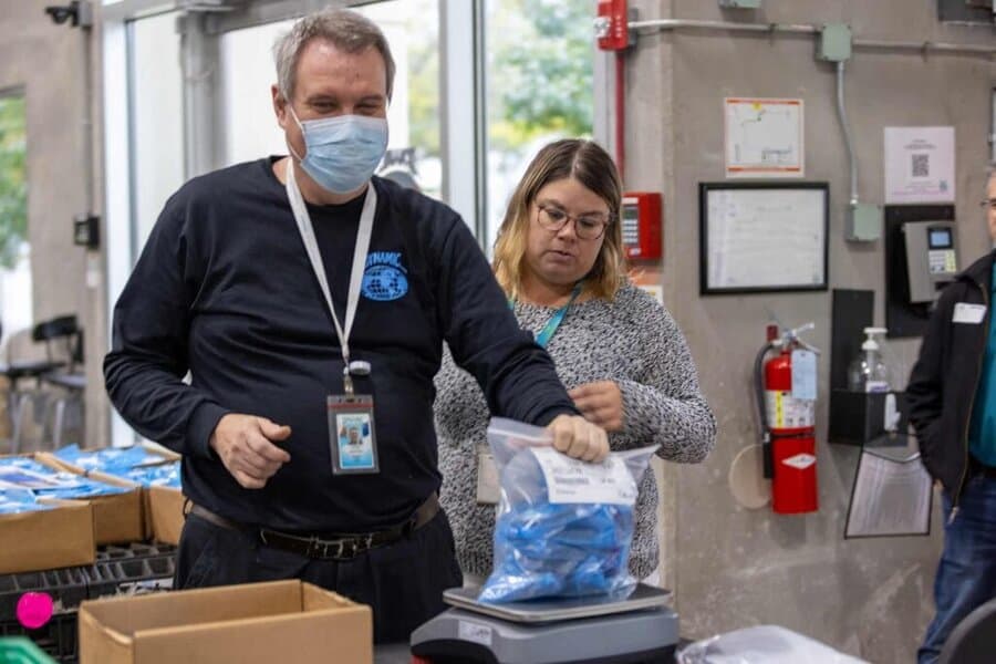 A man wearing a face mask places a sealed bag on a scale while a woman observes beside him. They are in an indoor setting with boxes, fire extinguisher, and safety equipment visible in the background.