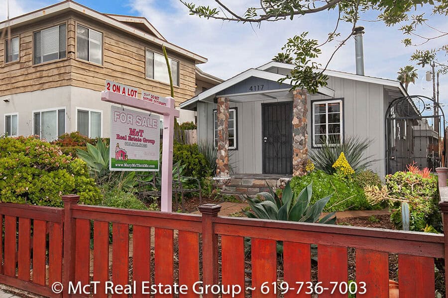 Exterior view of a North Park home on 33rd Street in North Park, showcasing a charming front house with unique stone pillars and a low wooden fence. This "2 on a lot" property is framed by lush, drought-tolerant landscaping and features a prominent McT Real Estate Group for-sale sign.