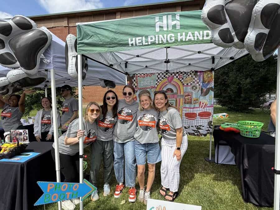 A group of five smiling women in matching "Helping Hands" shirts pose under a green canopy at an outdoor event, with a colorful backdrop and "Photo Booth" sign in the foreground.