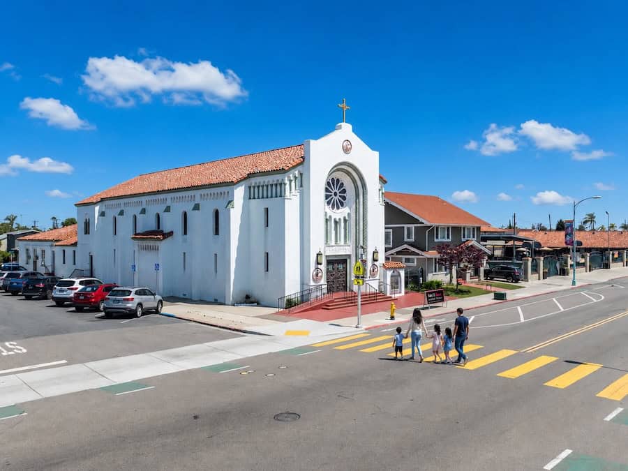 Parents and children walk across the crosswalk toward St. Patrick’s School in North Park, San Diego, on a sunny day with parked cars and a blue sky