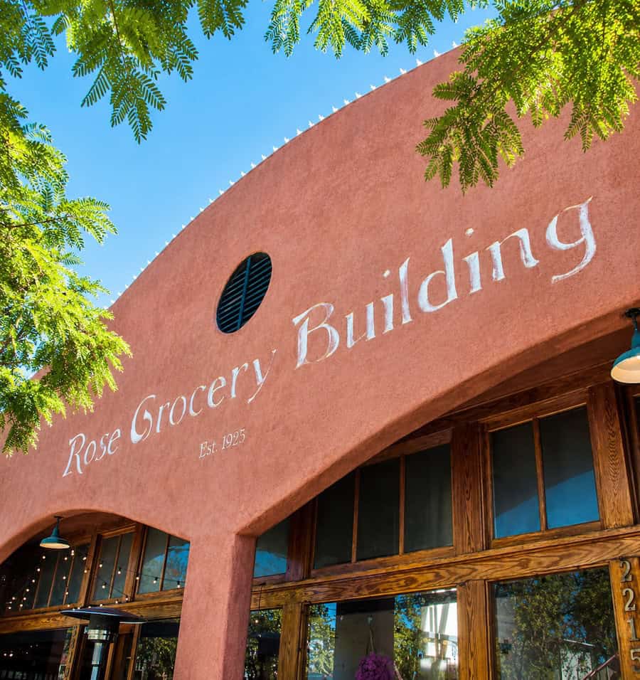 Rose Grocery Building exterior with warm stucco facade, wooden windows, blue sky, and leafy green tree branches framing the storefront.