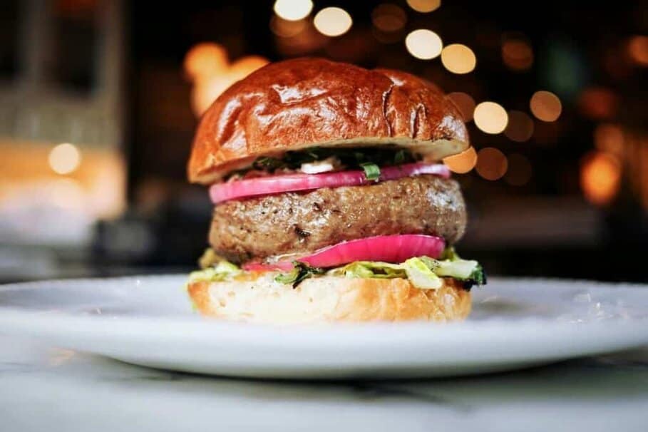 A close-up of a gourmet burger with a glistening brioche bun, thick beef patty, red onion rings and fresh greens, served on a white plate. The background features a bokeh effect with warm, twinkling lights, giving a cosy and inviting ambiance to the meal presentation for the best dinner in Blackpool.