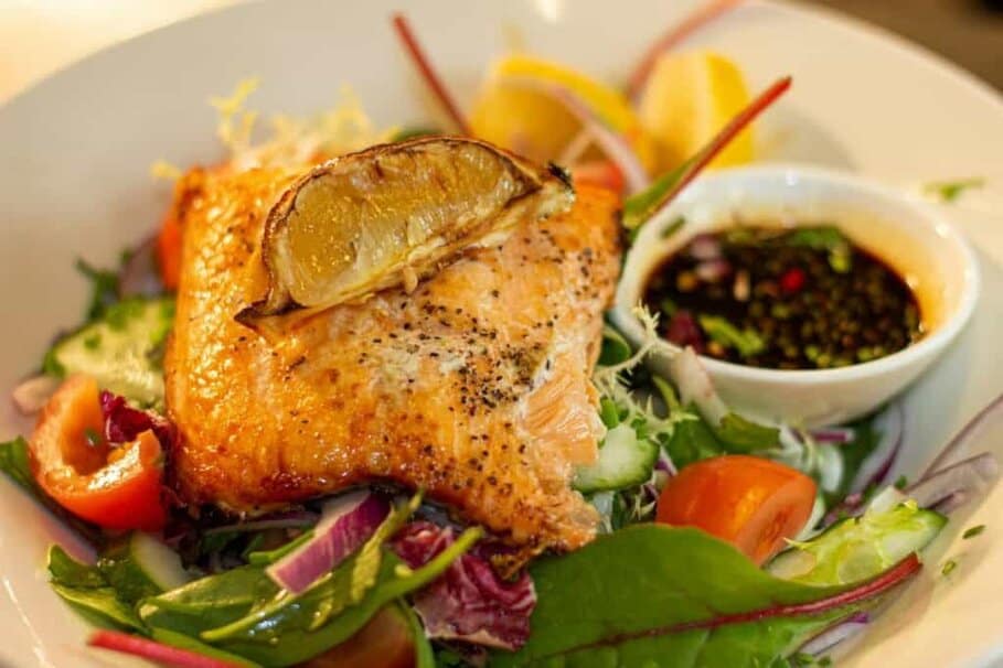 A succulent grilled salmon fillet topped with a grilled lemon slice, served on a fresh mixed greens salad with tomato and red onion, accompanied by a side of soy sauce dressing, presented as a feature dish for the best dinner in Bolton.