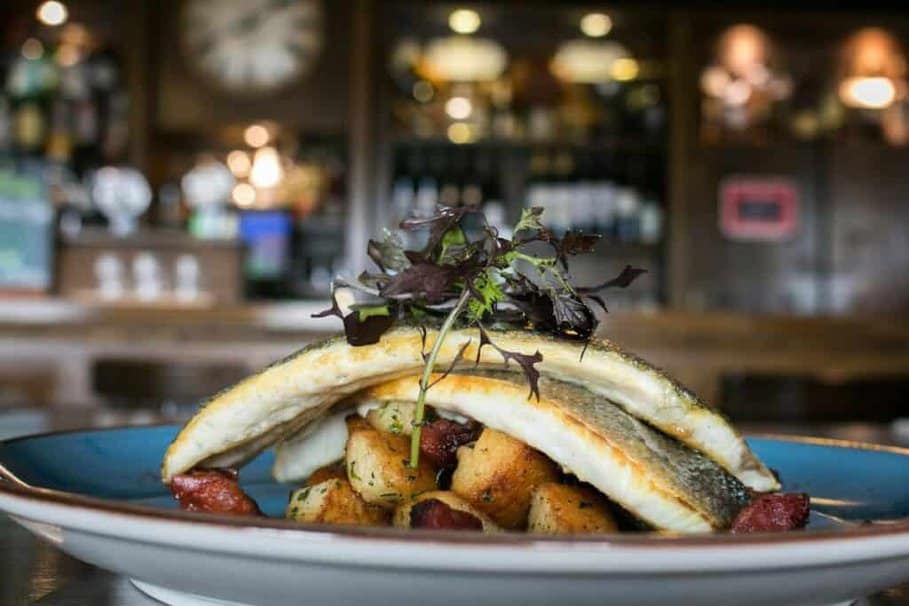 A freshly prepared dinner plate featuring grilled fish fillets with crispy skin, served over a bed of golden roasted potatoes, garnished with delicate mixed greens, presented on a blue ceramic plate with a blurred restaurant bar background, associated with the Best Dinner In Chester.