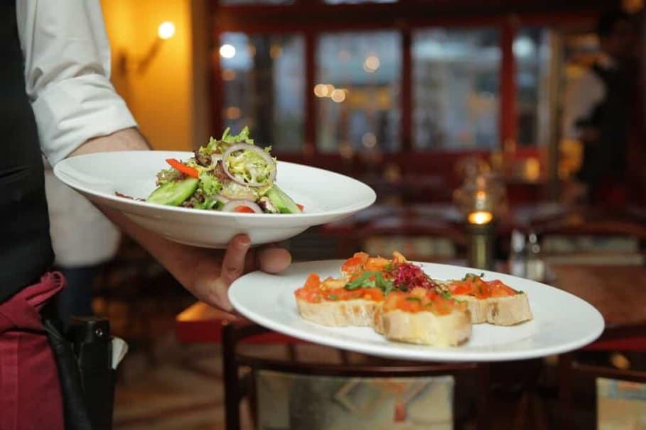Waiter serving a fresh salad and bruschetta on seperate white plates, in a restaurant, representing the best dinner options in Milton Keynes.