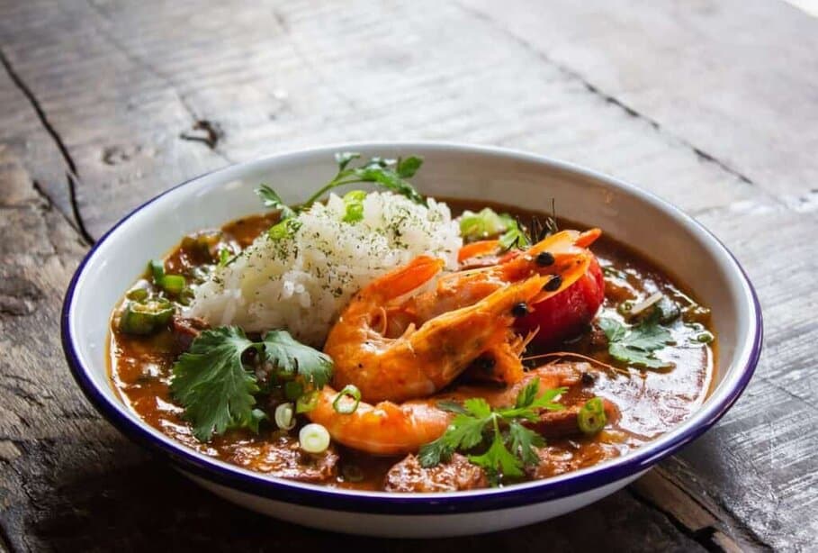 A bowl of shrimp gumbo with a side of rice garnished with herbs, presented on a rustic wooden table. The gumbo has a rich, savoury broth with shrimp, vegetables and seasoning visible, evoking a hearty, traditional meal, highlighting the best dinner in Blackburn.