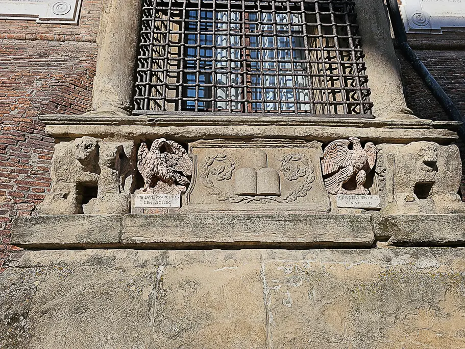 Detailed stone reliefs featuring eagles and classical symbols under a window of Palazzo d’Accursio, showcasing Bologna’s heraldic art and Renaissance design elements.