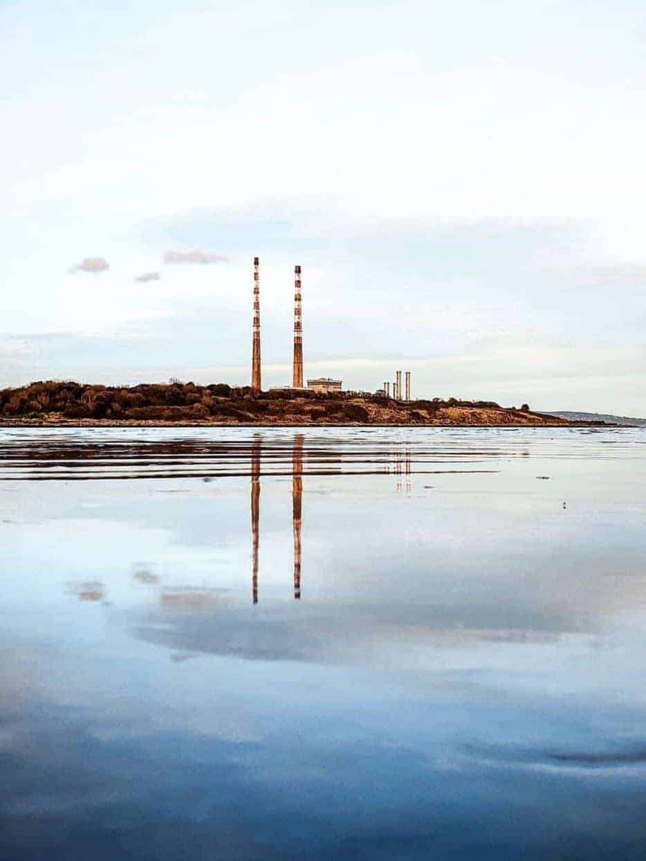 A view of the Poolbeg Chimneys in Dublin, with the twin red and white towers reflected in the calm water of Dublin Bay, under a pale sky.