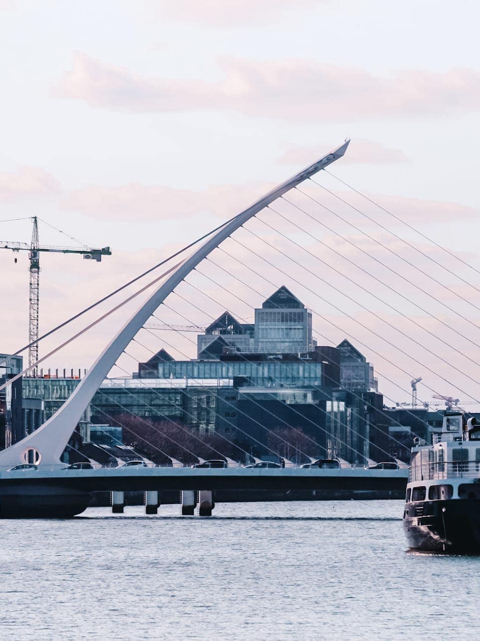 The Samuel Beckett Bridge in Dublin, designed to resemble a harp, with modern office buildings and construction cranes in the background, viewed over the River Liffey.