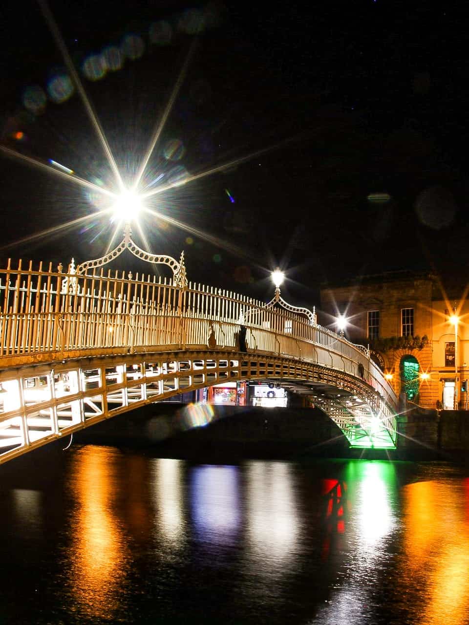 The Ha'penny Bridge in Dublin at night, with bright lights illuminating the bridge and their reflections creating a colorful display on the river's surface.