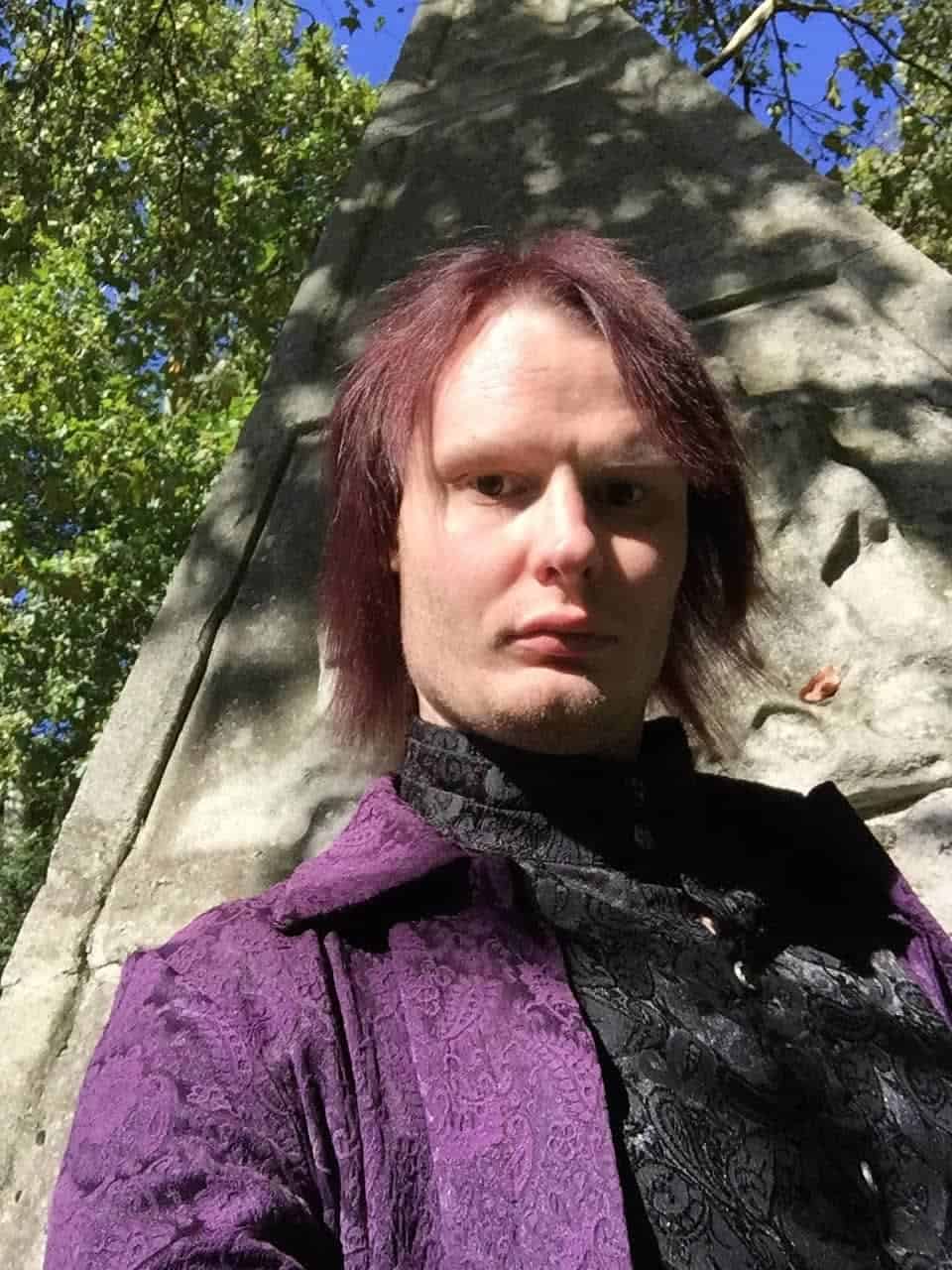 Mid-length portrait of a person with reddish-brown hair standing outdoors against a large stone monument, surrounded by green trees, under a bright blue sky, wearing a purple and black patterned jacket.
