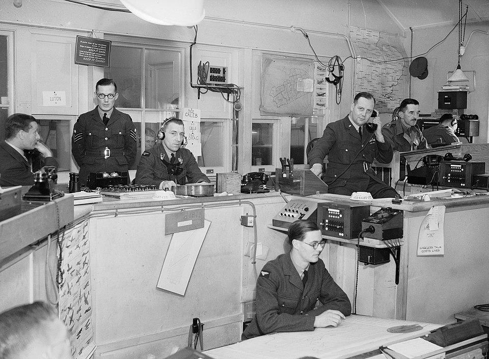 Operatiekamer Sector G, Duxford 1940: RAF-personeel coördineert jachtvliegtuigen met kaarten, radio en squadron-codes op de muur.