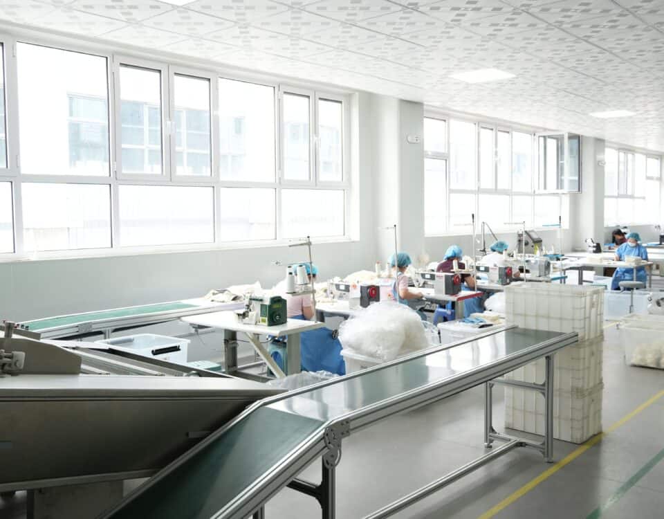 Workers wearing protective headwear operate assembly workstations along a conveyor belt in a bright manufacturing facility.